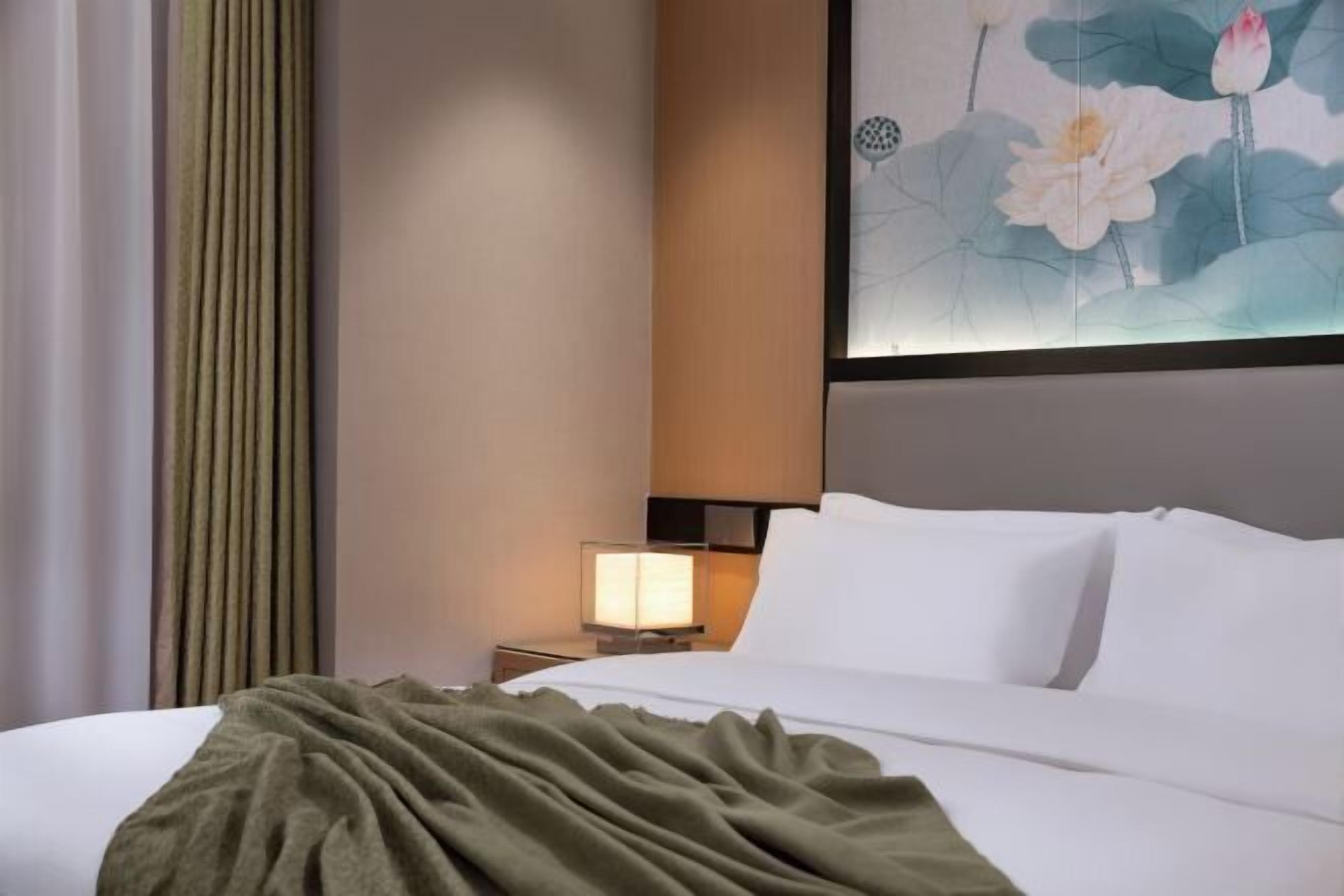 Habitación de hotel con una cama bien hecha, una mesita de noche con lámpara y un cuadro de flores de loto en la pared.