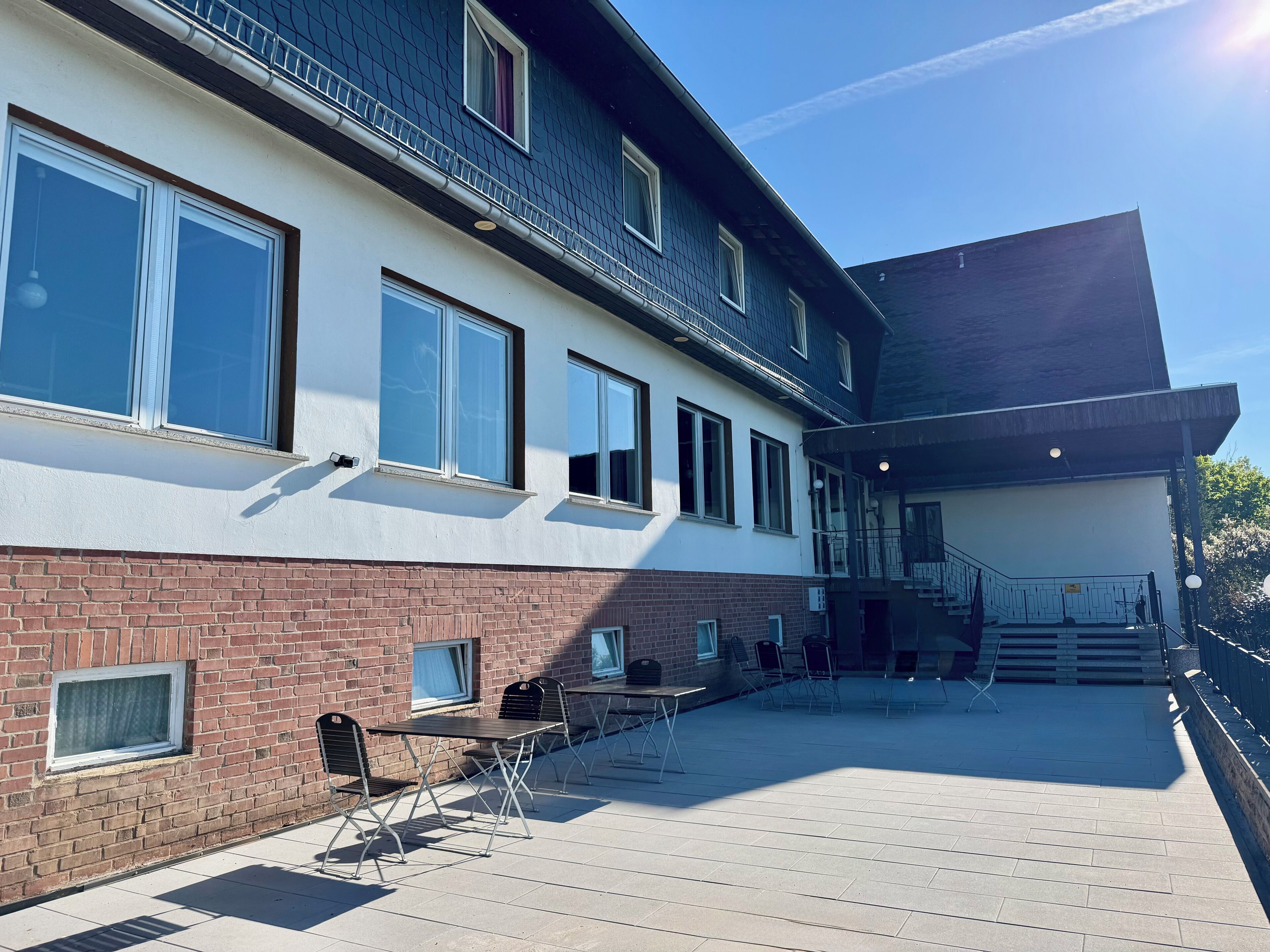 En byggnad med en nedre del i tegel och en vit övre del, med flera fönster och ett takförsett utomhusområde med bord och stolar.
