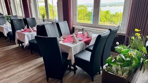 A restaurant with tables set for dining, large windows with a view of greenery and water, and a potted plant on the right. - Barbarossa Hotel Kelbra (Kelbra)