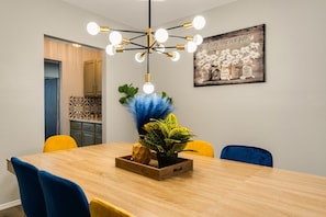A dining area with a wooden table, blue and yellow chairs, a decorative plant, and a wall-mounted light fixture. - Comfortable 5BR 3BA • Central Location (Rowlett)