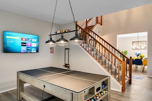 A ping pong table in a room with a TV displaying streaming services, a staircase, and a dining area. - Comfortable 5BR 3BA • Central Location (Rowlett)
