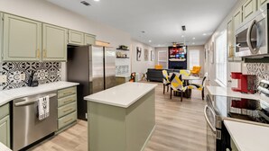 A modern kitchen with green cabinets, stainless steel appliances, and a central island. There is a dining area with yellow chairs and a fireplace in the background. - Comfortable 5BR 3BA • Central Location (Rowlett)