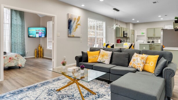 A modern living room with a grey sofa, yellow pillows, and a glass coffee table. - Comfortable 5BR 3BA • Central Location (Rowlett)