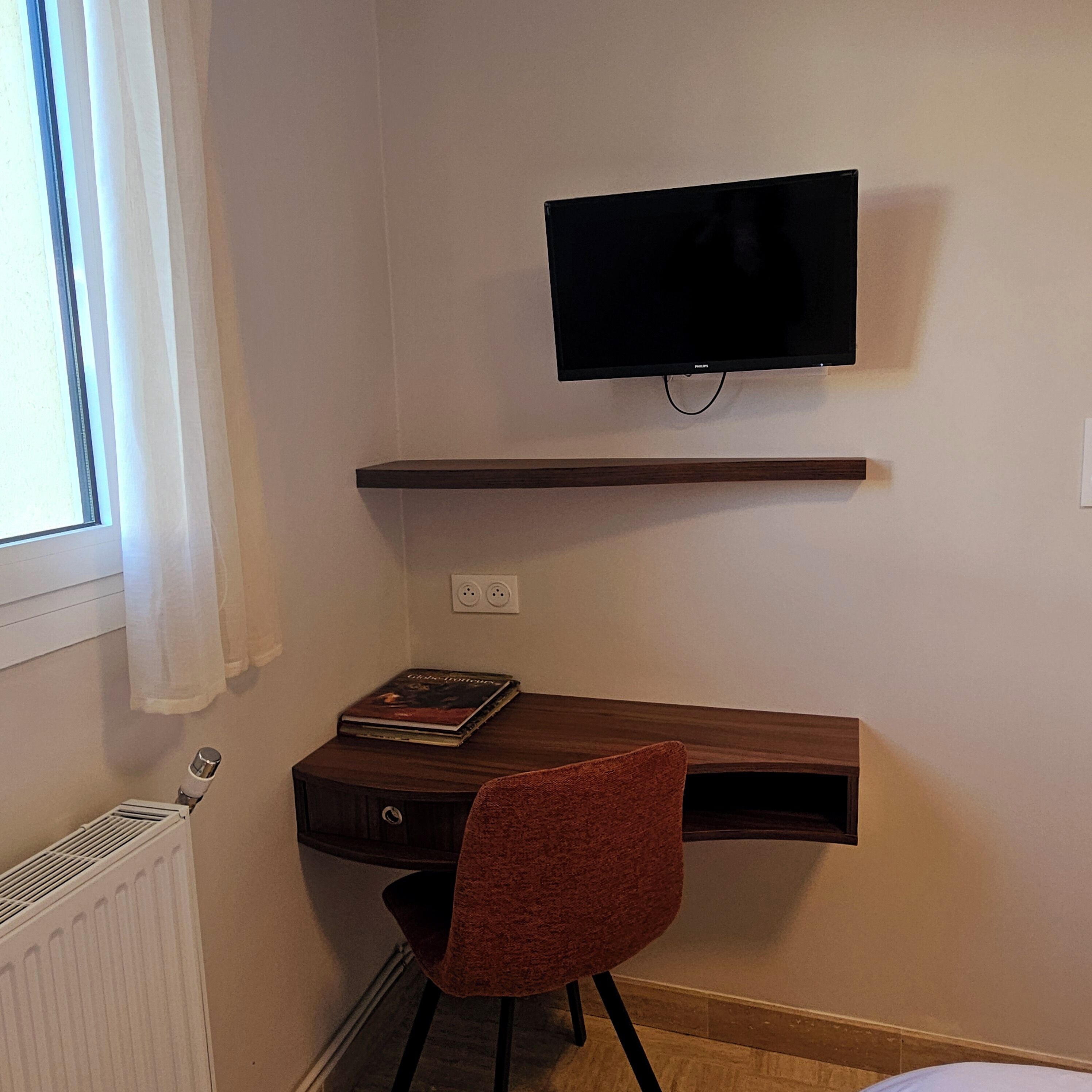 A small room with a desk, a chair, a television mounted on the wall, and a window with curtains.