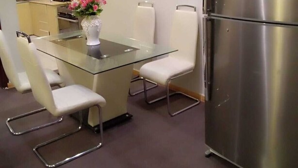 Una cocina moderna con un refrigerador de acero inoxidable, una mesa de vidrio con sillas blancas y un florero con flores.