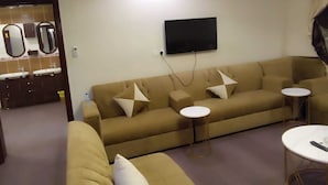 Una sala de estar con un sofá, dos mesitas auxiliares, un televisor de pantalla plana montado en la pared y un baño con un espejo y dos lavamanos.