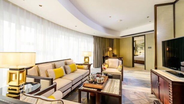 A hotel room with a sofa, armchair, coffee table, TV, and a large window with curtains. - Dahe Jinjiang Hotel (Zhengzhou)