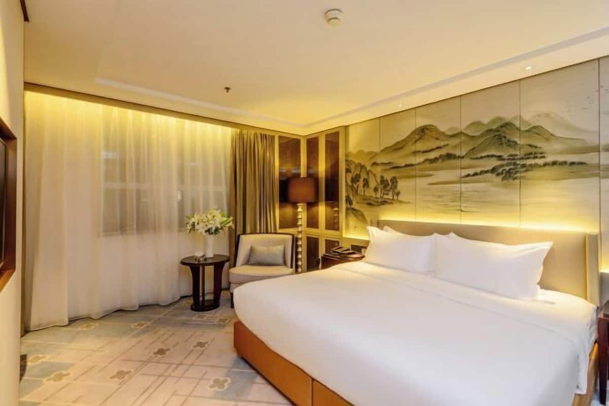 Hotelkamer met een groot bed, nachtkastje, stoel, een klein tafeltje met een bloevas en een schilderij aan de muur.