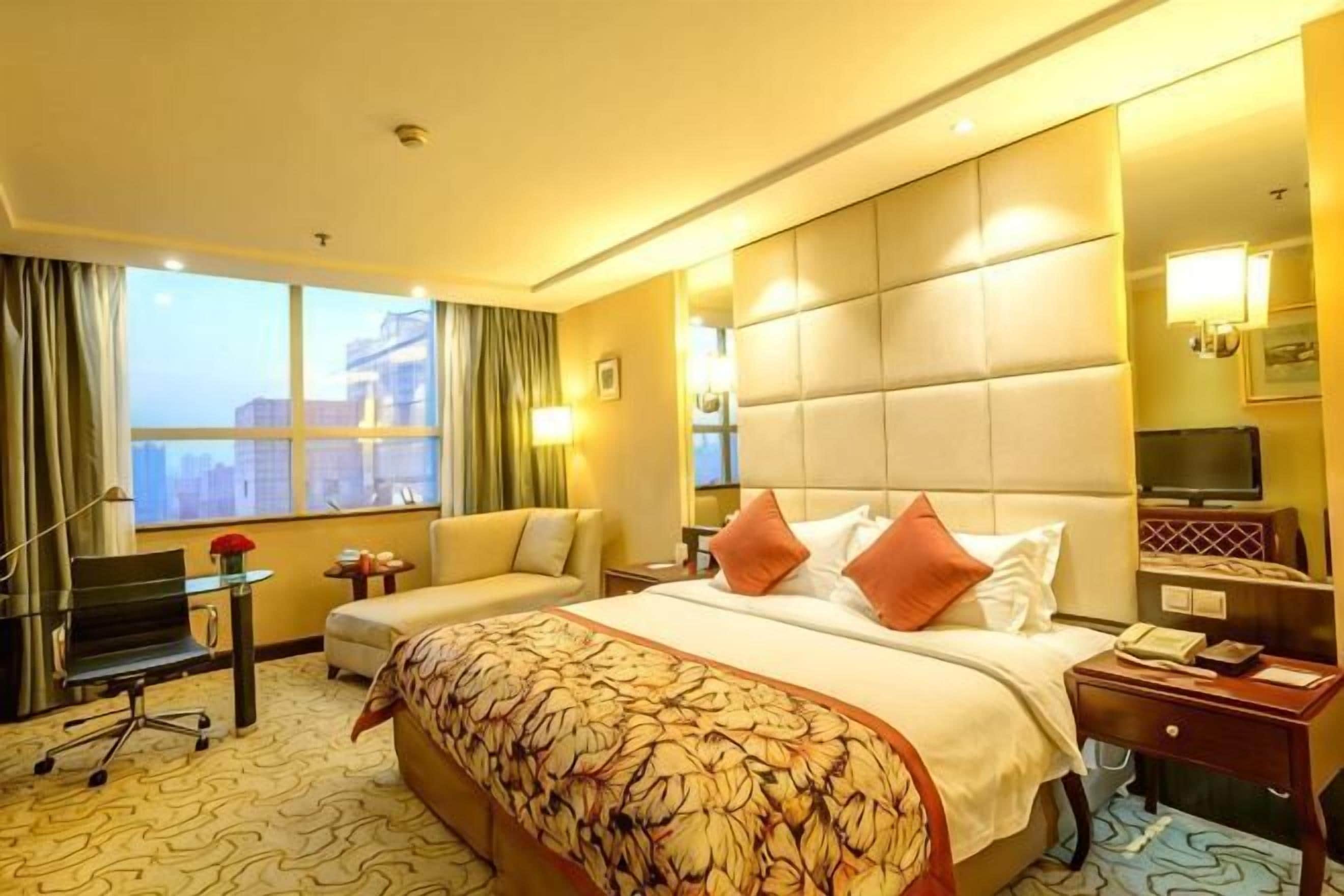 A hotel room with a large bed, a desk, a chair, a TV, and a view of the city.
