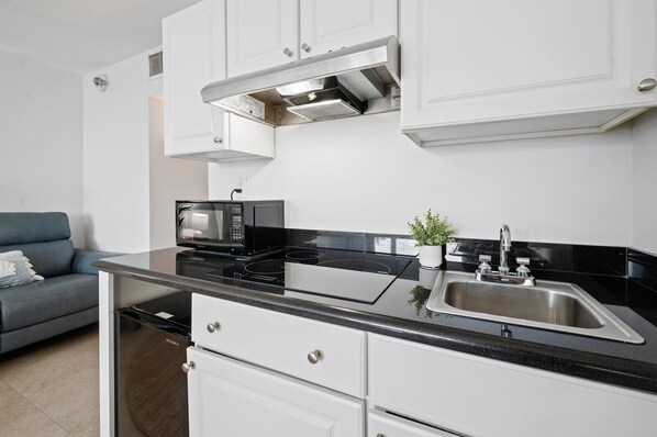 A compact kitchen with white cabinets, a black countertop, a built-in microwave, a stainless steel sink, and a compact dishwasher.