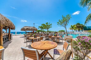 An outdoor dining area with round wooden tables and chairs, a pool, and a view of the ocean. - Ocean Manor 1BR - Beachfront, Pool & Tiki Bar (Fort Lauderdale)