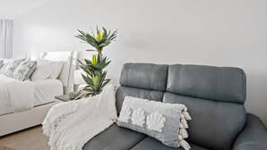 Quarto moderno com um sofá cinza, uma cama branca com almofadas estampadas e um vaso de planta.