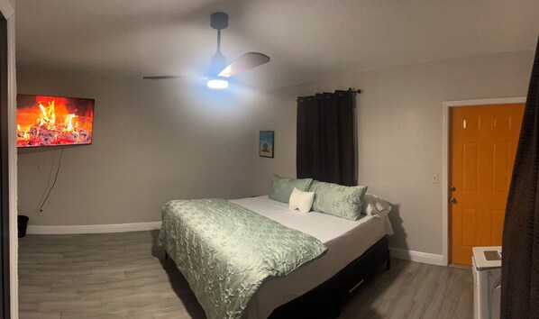 Une chambre à coucher avec un lit, un ventilateur de plafond, un téléviseur diffusant une image de feu et une porte.