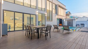A modern outdoor deck with a pool, a dining area with chairs, and a large glass door leading to a living space.