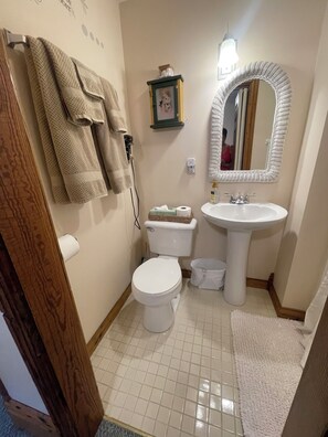 A bathroom with a toilet, sink, and a mirror. There is a towel rack with towels, a small shelf with toiletries, and a trash bin.