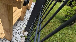 Un banco de madera con estructura metálica, situado en un camino de grava con una valla metálica al fondo.