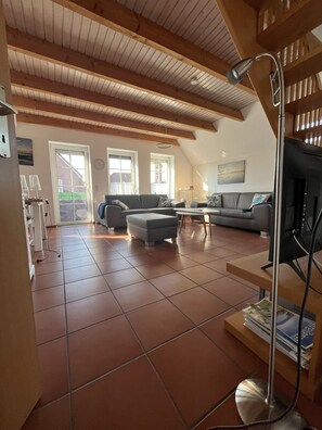 A spacious living area with a high ceiling, tiled floor, and a television on a stand. - Detached vacation home in Nessmersiel (near Deich) with private garden & wallbox (Dornum)