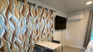 A room with a dining table, a chair, a TV mounted on the wall, and an air conditioning unit.