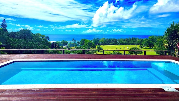 Ein Schwimmbecken mit Holzterrasse, umgeben von üppiger Vegetation und Blick auf das Meer unter blauem Himmel.