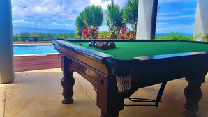 Ein Billardtisch mit grüner Spielvliesbahn, umgeben von einem Betonboden, unter einem überdachten Bereich mit Blick auf einen Swimmingpool und tropische Pflanzen.