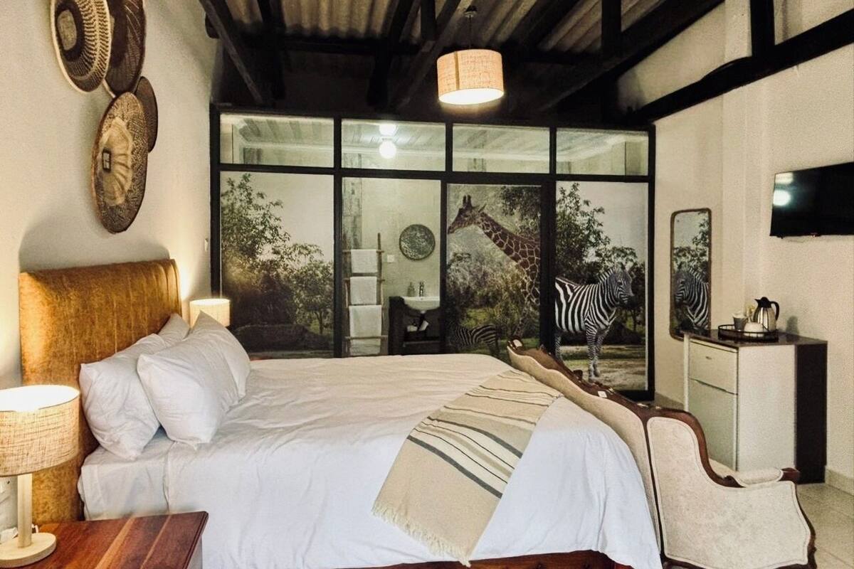A bedroom with a large bed, a wooden headboard, a bedside table with a lamp, a dresser, and a large window with a view of a giraffe and zebra.