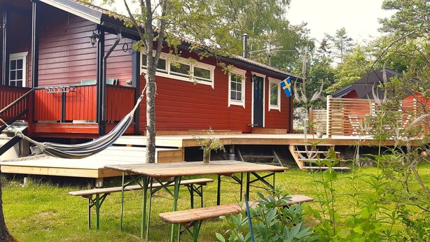 A red wooden cabin with a hammock, picnic table, and Swedish flag. - Nice, comfortable vacation home 4 beds, with terrace, near the lake (Stockholms län)