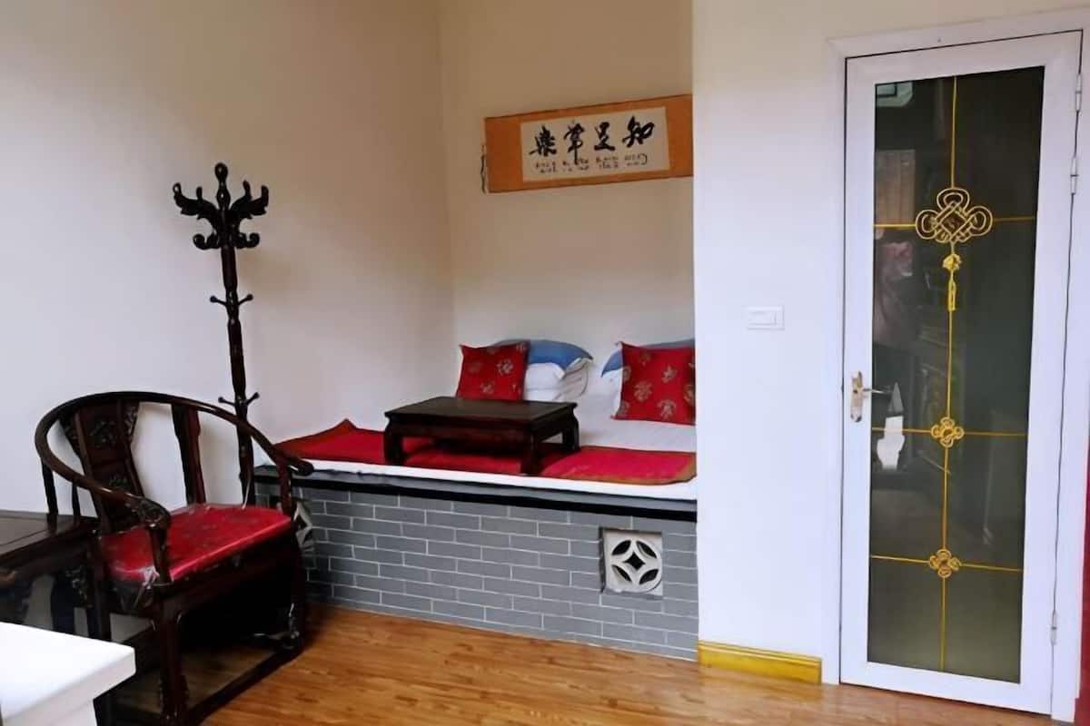 A room with a wooden floor, a sofa with red cushions, a wooden chair, a coat rack, and a door with a decorative design.