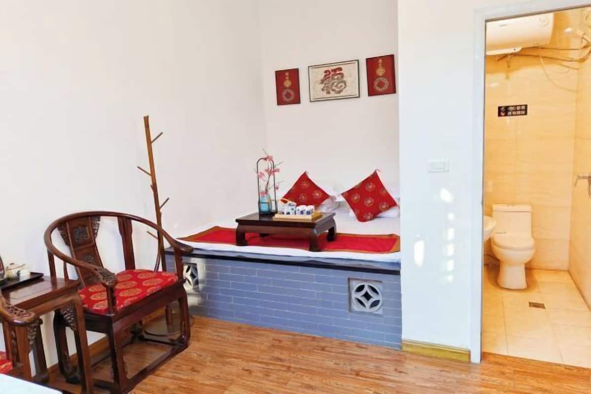 A room with a bed, a wooden chair, a small table with a lamp, and a toilet.