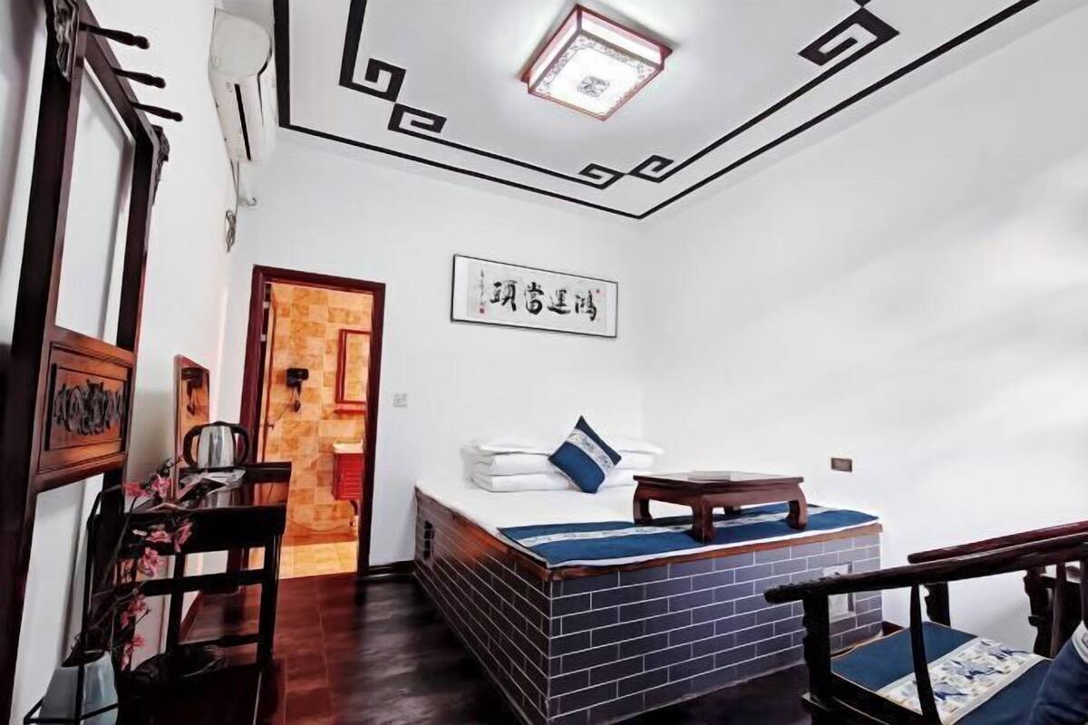 A room with a bed, a small table, a chair, a wall-mounted shelf, and a door.
