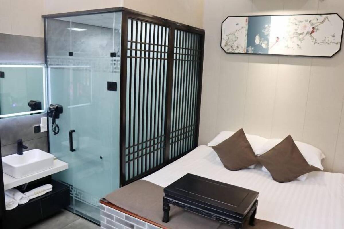 A hotel room with a glass-enclosed shower, a white sink, a bed with brown pillows, and a small table.