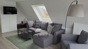 A modern living room with a grey sofa set, a glass coffee table, a lamp, and a TV mounted on the wall.