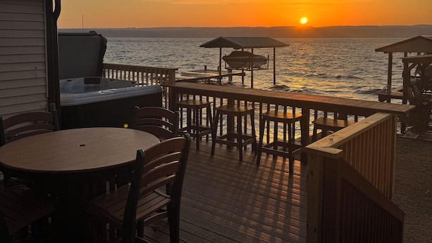 A wooden deck with a table and chairs, overlooking a body of water at sunset.