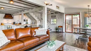 A spacious living area with a brown leather sofa, a coffee table, and a dining table with chairs. There is a staircase leading to an upper level, and a kitchen area is visible in the background.