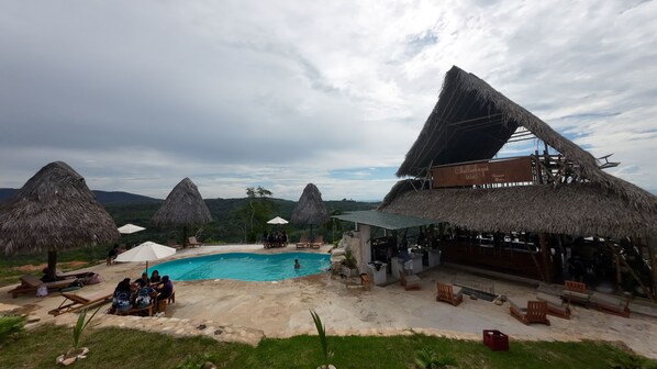 A poolside area with thatched-roof structures, lounge chairs, and a bar named 'Chullachagui Wasi'. - Renaco Glamping (Sauce)