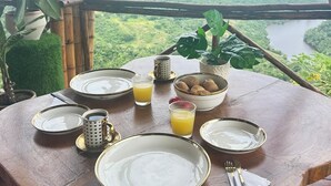 Une table de salle à manger en bois, mise avec des assiettes, des tasses et un saladier rempli de fruits, donnant sur un paysage luxuriant avec une rivière.