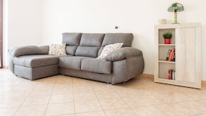 A grey sectional sofa with floral cushions, a white bookshelf with books and a small plant, and a lamp on top. - Villa Vespucci - Pet Friendly (Montesilvano)
