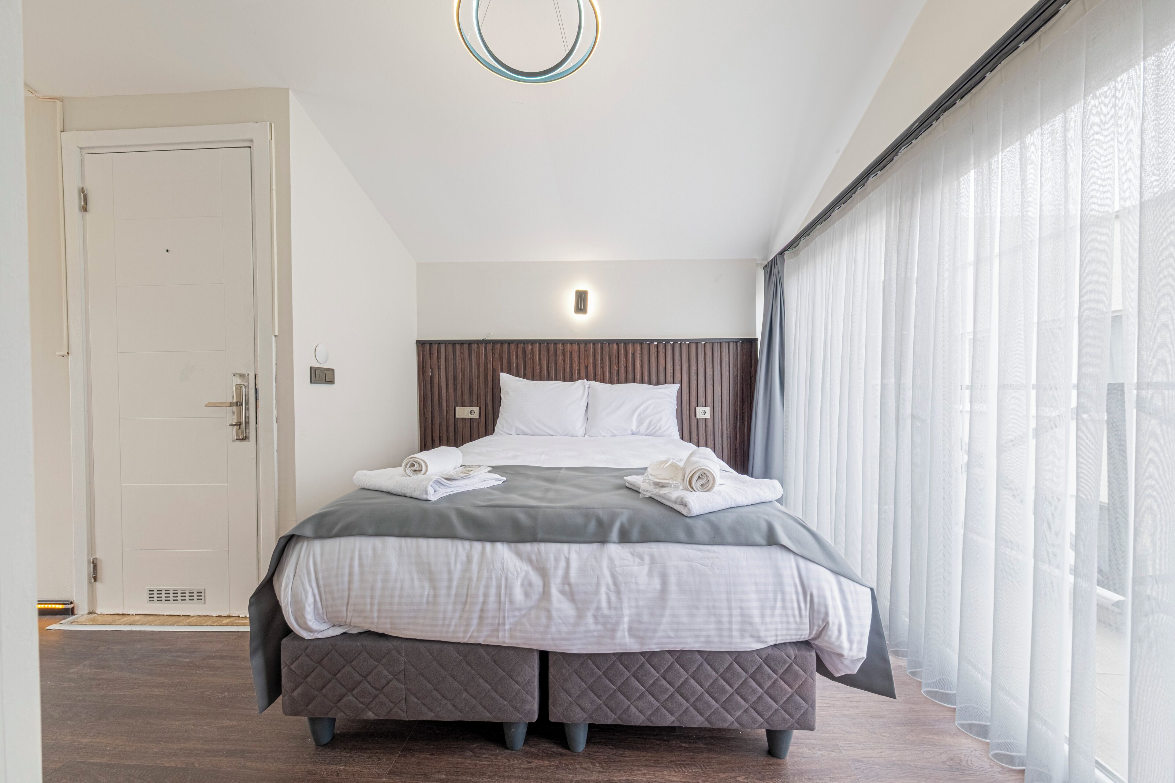 Camera d'albergo moderna con un letto grande, una testiera in legno, un comodino e una finestra con tende leggere.