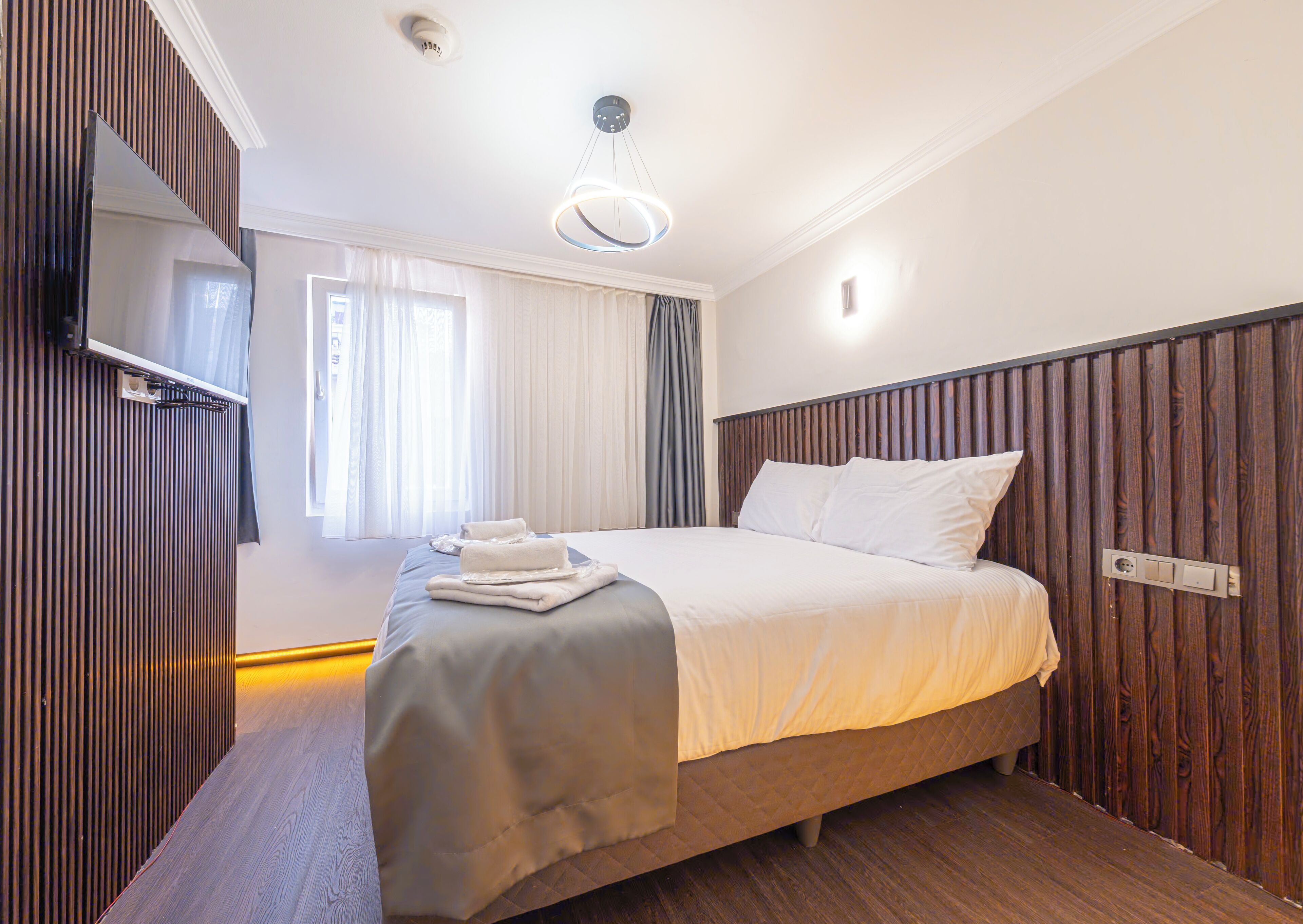 A modern hotel room with a large bed, wooden paneling on the walls, a flat-screen TV, and a window with curtains.