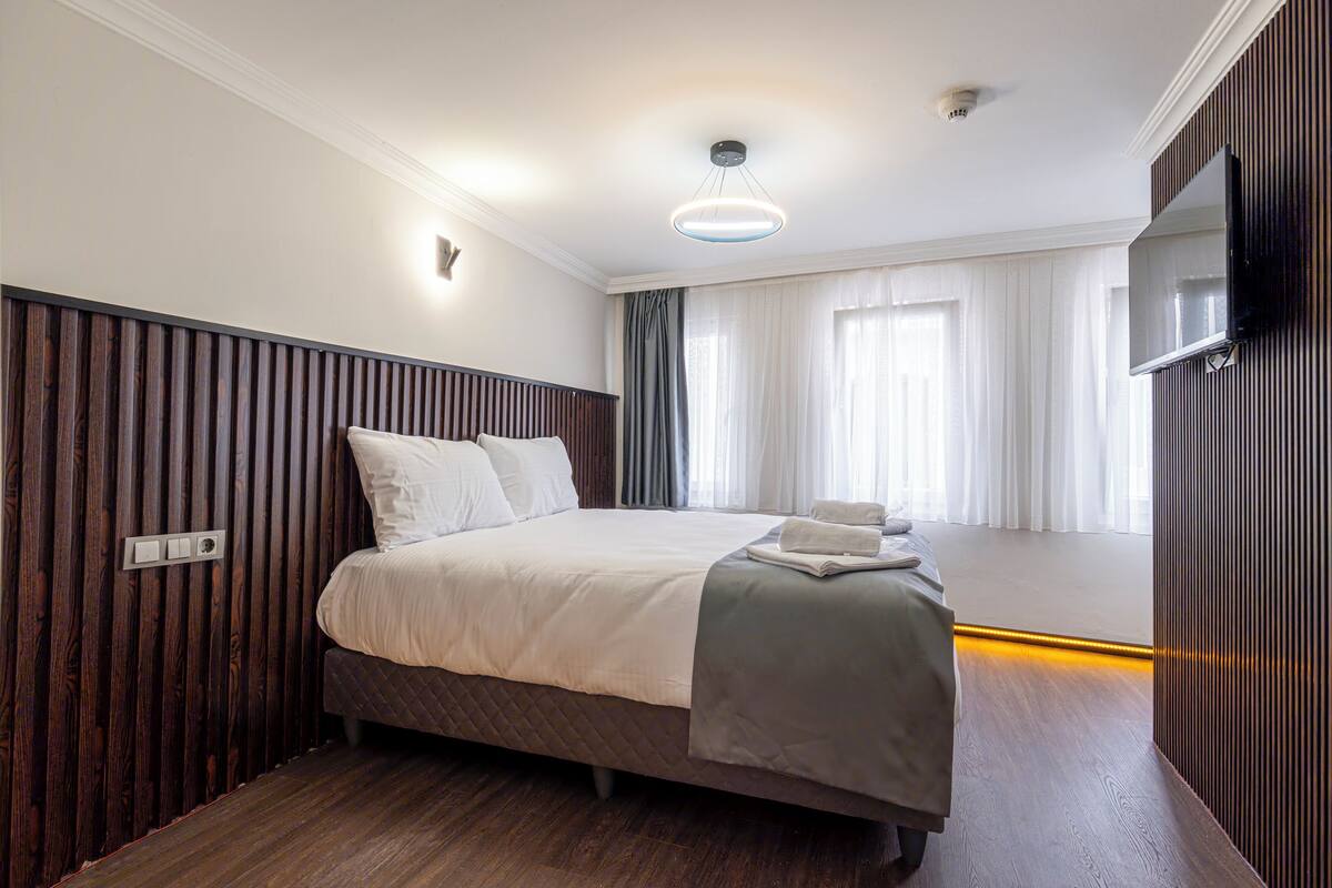 A modern hotel room with a large bed, a flat-screen TV mounted on the wall, and a large window with curtains.