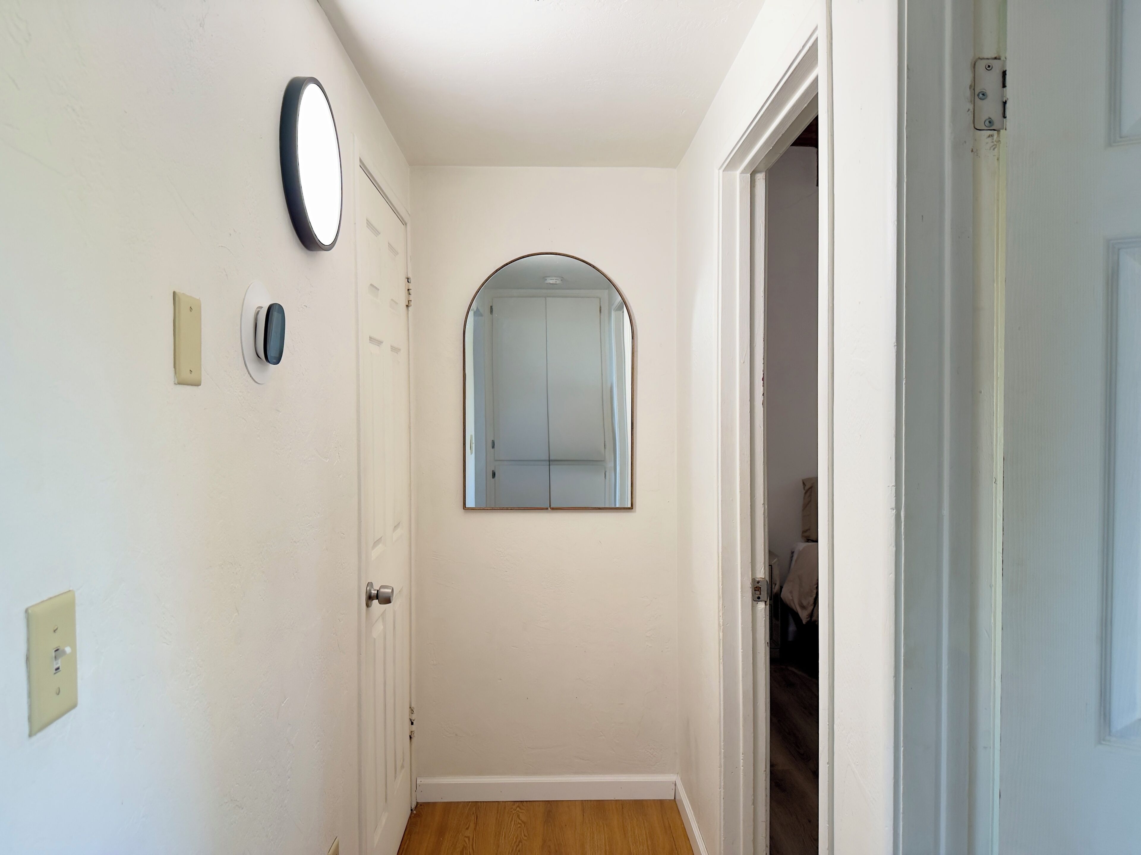 A narrow hallway with a white door, a wall-mounted round mirror, and a doorbell.