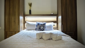 Una cama bien hecha con ropa blanca y dos almohadas, cabecera de madera y un florero con plantas secas en la pared.