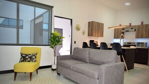 Una sala de estar moderna con un sofá, un sillón amarillo y un comedor con sillas, y una cocina al fondo.