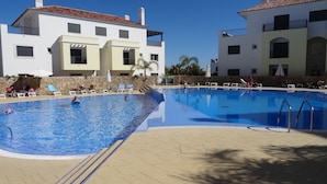 A swimming pool with lounge chairs and umbrellas, surrounded by buildings with balconies. - The Pomar 2 Bedroom Suite -5 People Terrace Swimming Pools (Tavira)