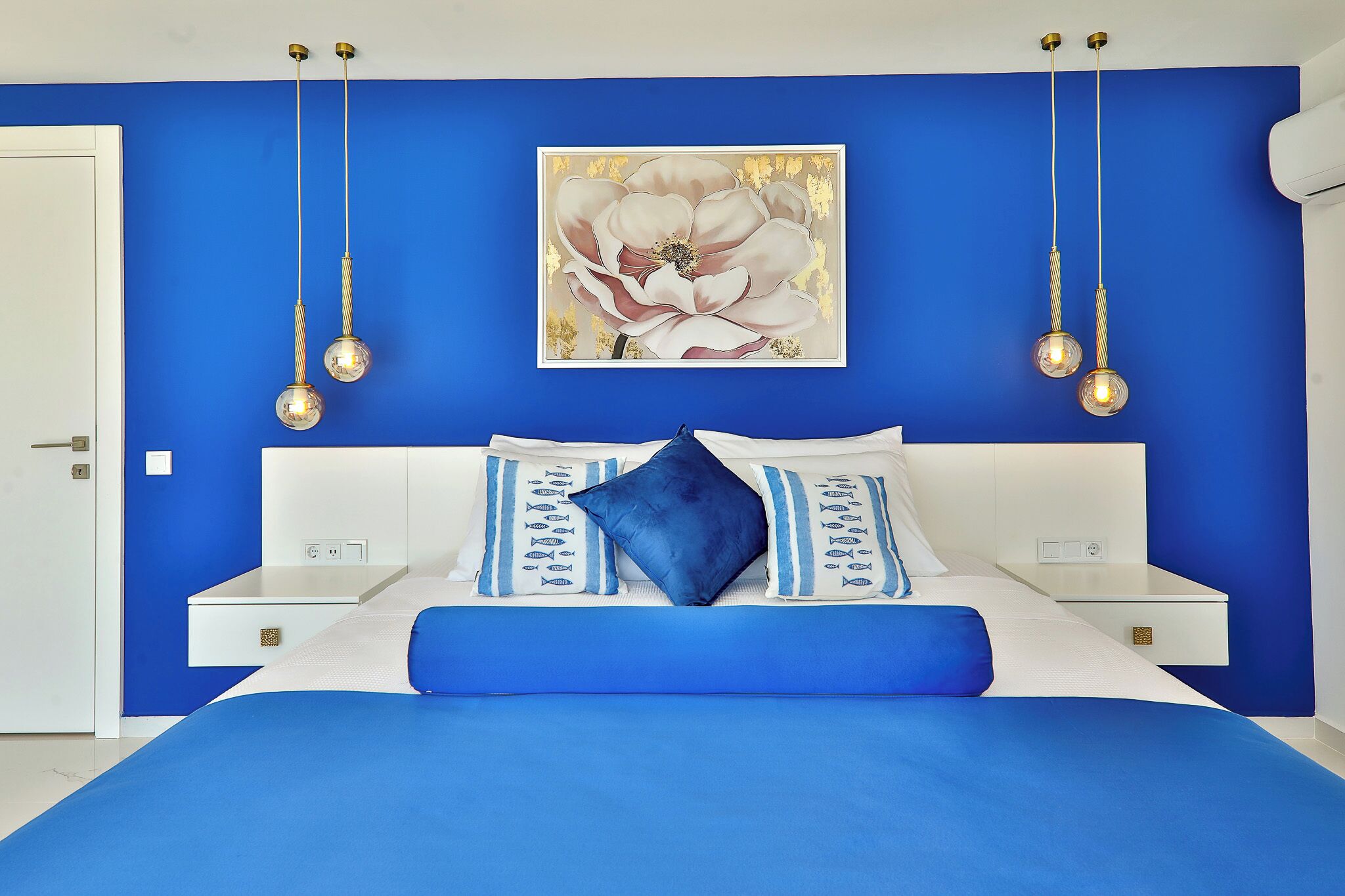 Un dormitorio con una paleta de colores azul y blanco, una cama grande con ropa de cama azul, dos mesitas de noche y un aplique de pared.