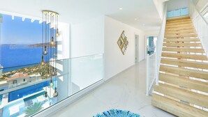 Un interior moderno con balcón de vidrio que da a una piscina y vistas al mar, una escalera de madera y una obra de arte geométrica en la pared.
