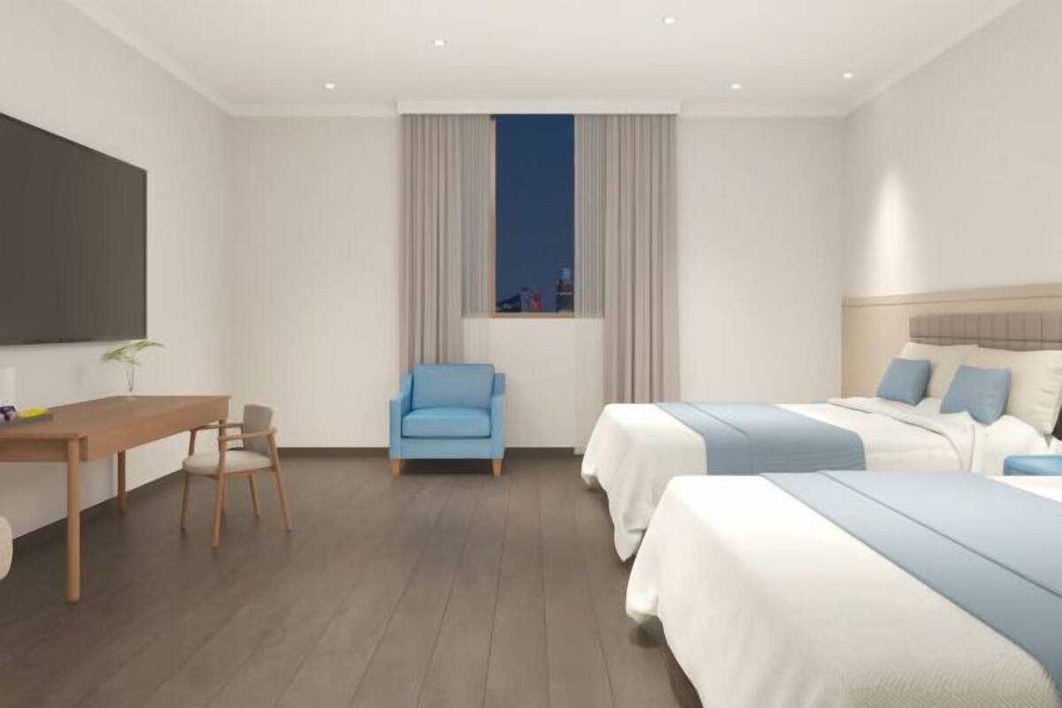Una habitación de hotel moderna con un escritorio de madera, un sillón azul, dos camas con ropa de cama azul, un ventanal con cortinas y un televisor de pantalla plana montado en la pared.