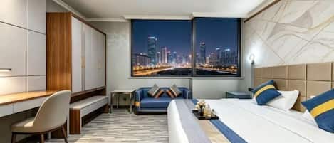 A hotel room with a bed, desk, chair, and a view of a cityscape at night.