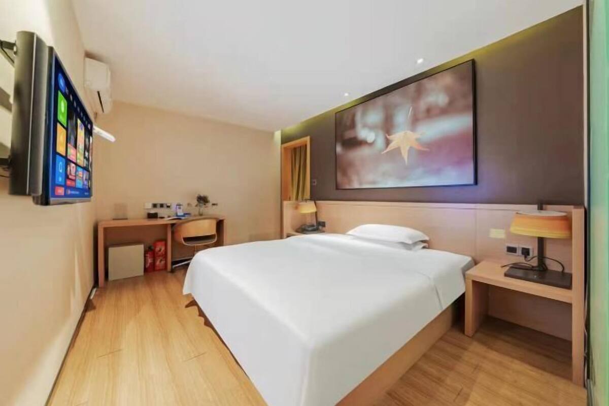 Una habitación de hotel con cama, escritorio, silla, televisor montado en la pared y un cuadro sobre la cama.