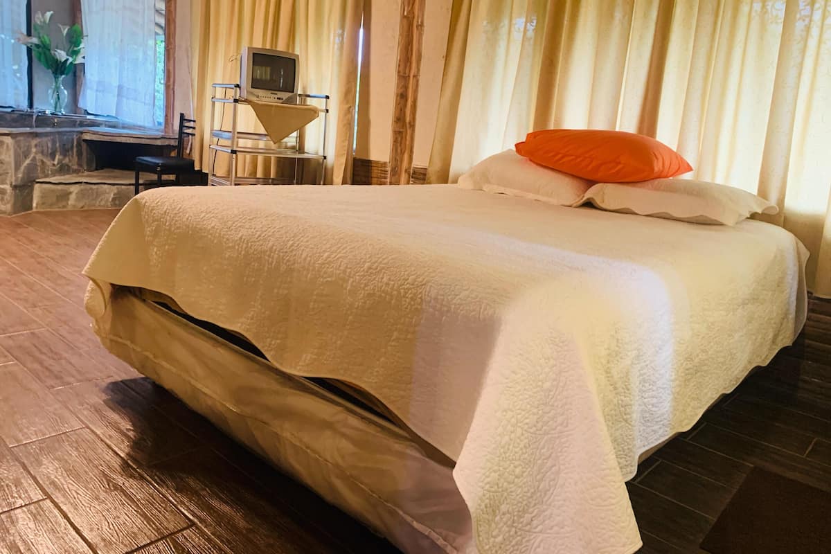 A neatly made bed with a white comforter and an orange pillow, a small desk with a computer, and a window with curtains.