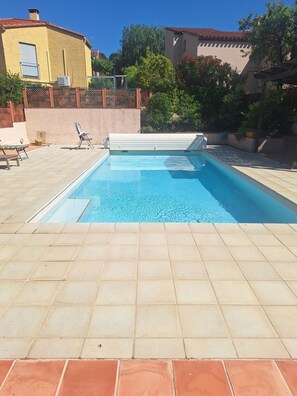 A swimming pool with a tiled deck, surrounded by a fence and trees.